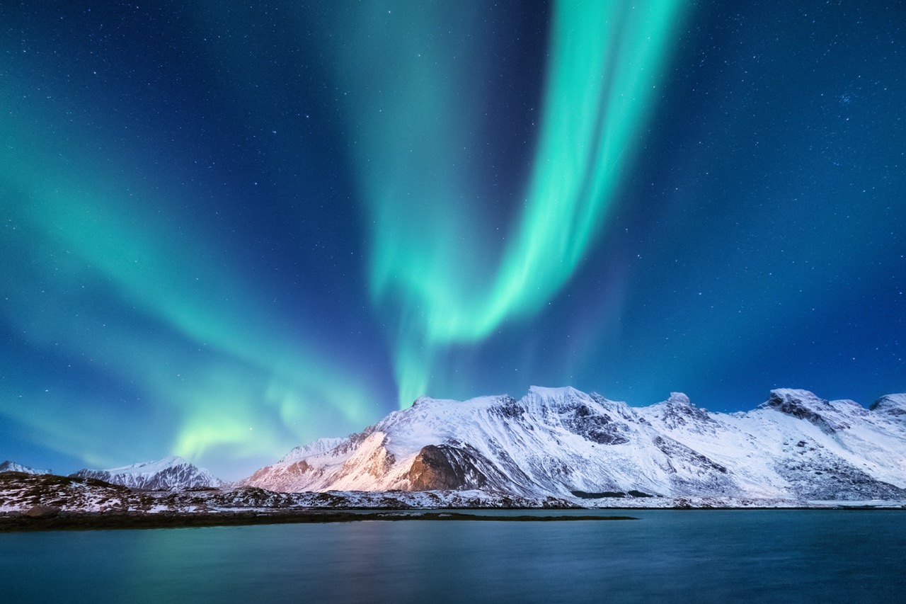 Spectacular aurora borealis dancing in vibrant green and blue hues above snow-covered mountains and a calm fjord waters. The dramatic night sky illuminates the rugged winter landscape with ethereal light.