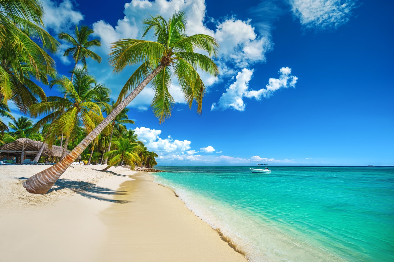A pristine white sandy beach with leaning palm trees overlooking crystal-clear turquoise waters with a small boat in the distance. Fluffy white clouds dot the vibrant blue sky, creating a picture-perfect tropical scene.