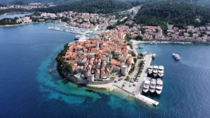 Aerial View of Rovinj Coastal Town An aerial photograph of the picturesque Rovinj, Croatia, showcasing the historic old town's terracotta-roofed buildings clustered on a peninsula surrounded by crystal-clear turquoise Adriatic waters, with several boats moored nearby and lush green forests visible in the background.