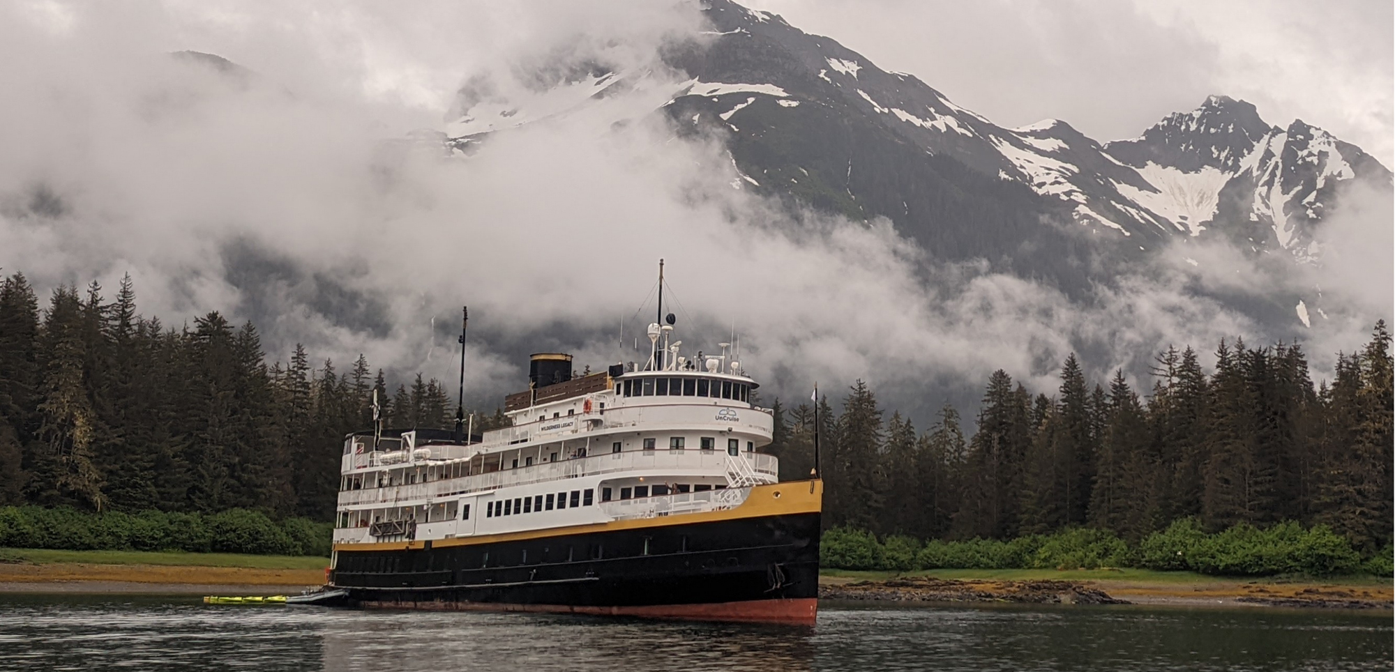 Alaskan Ferry in Misty Mountain Landscape A white and yellow passenger ferry navigates through calm waters near a forested shoreline with snow-capped mountains shrouded in fog in the background.