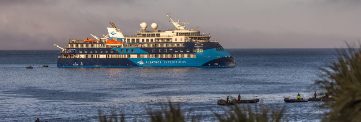 The Albatros Expeditions cruise ship is anchored in calm coastal waters, displaying its distinctive blue and black hull with multiple passenger decks. Small tender boats are visible in the foreground, suggesting passenger excursions to the nearby shore with beach grasses framing the scene.