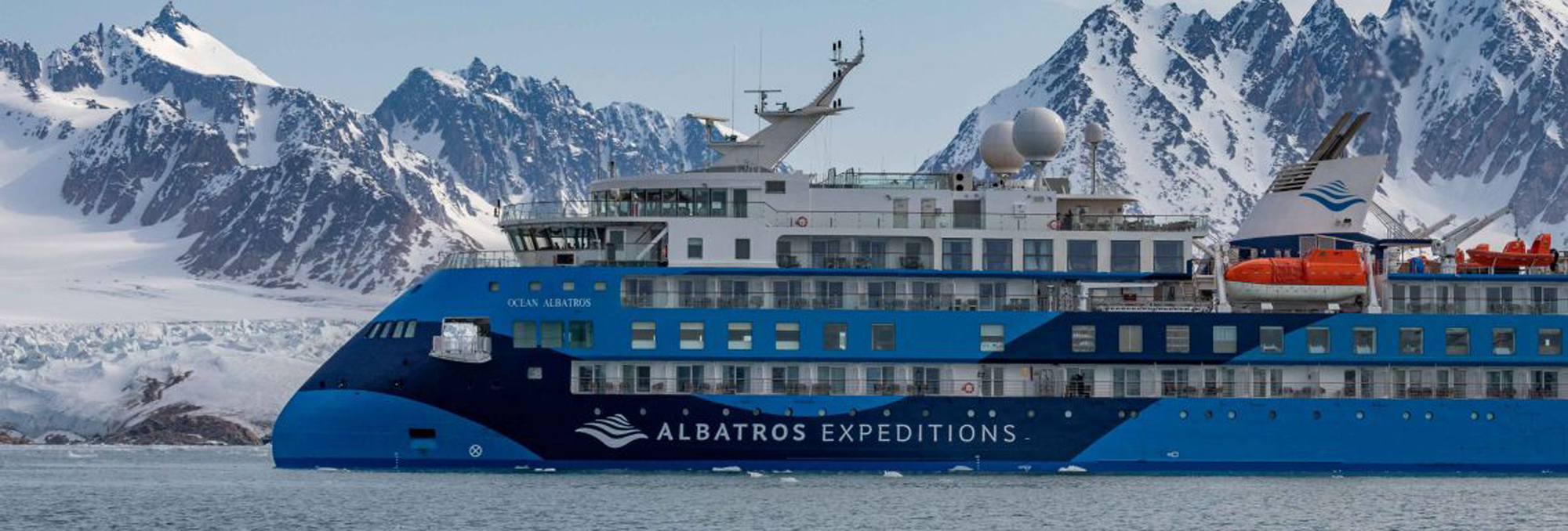 A blue and white expedition cruise ship navigating through icy waters with snow-capped mountains in the background. The Ocean Albatros vessel is designed for polar exploration, featuring rescue boats and a robust design.