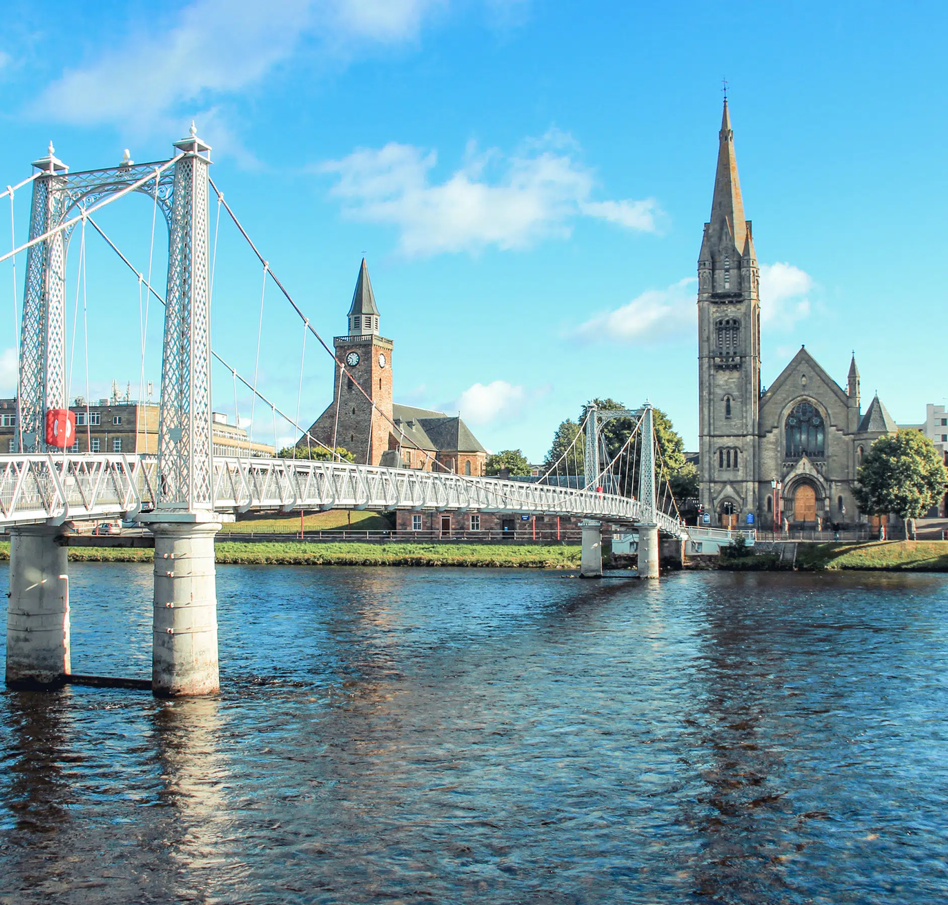 Albert Bridge and Churches in Inverness A picturesque view of the white ornamental Albert Bridge spanning the River Ness in Inverness, Scotland, with historic church spires visible in the background against a bright blue sky. The iconic cable-stayed bridge showcases Victorian engineering alongside traditional Scottish architecture.