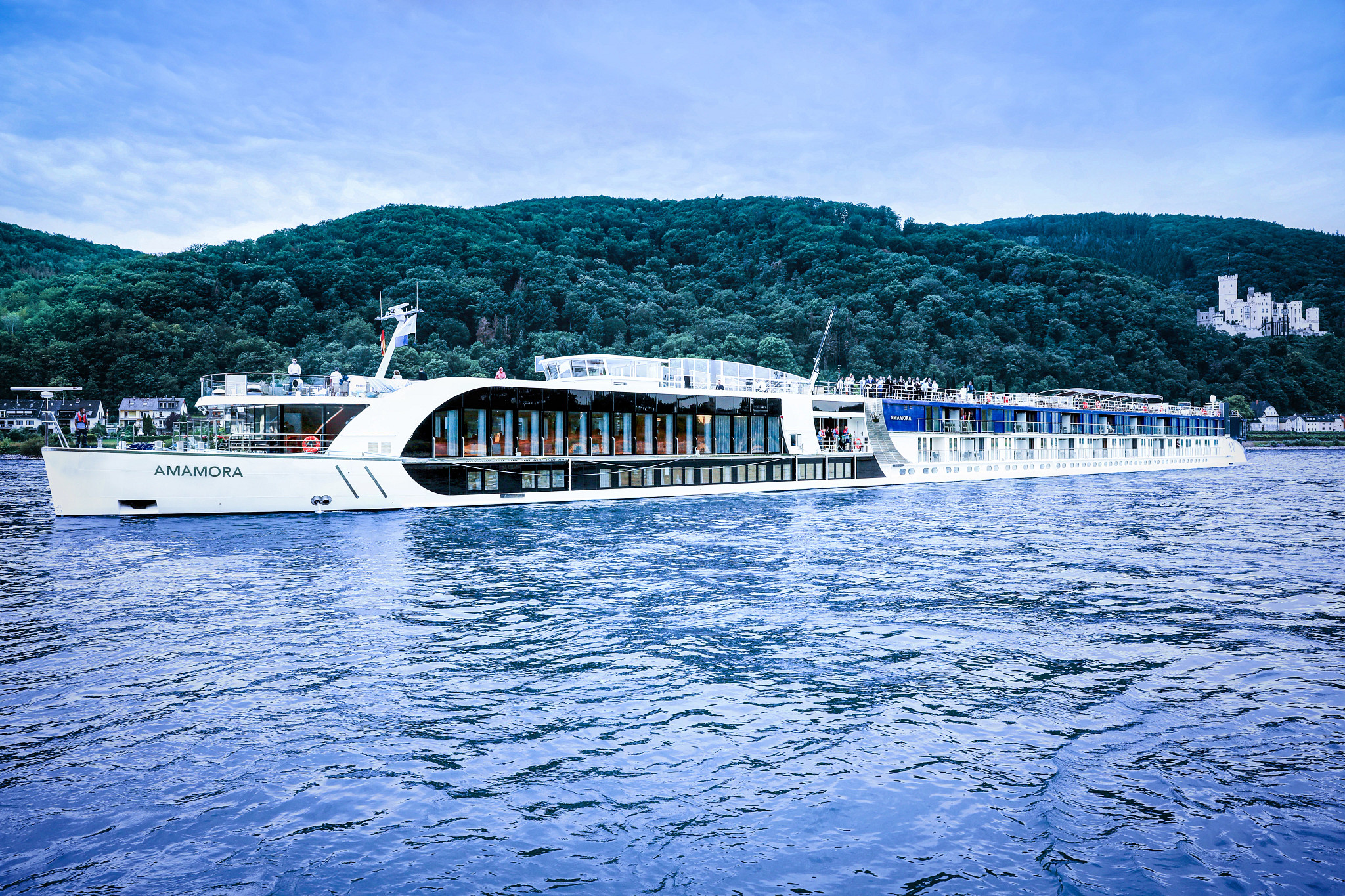 A modern river cruise ship named AmaMora sailing on a blue river with lush green forested hills and a white castle in the background. The vessel features large panoramic windows and a sleek white design.