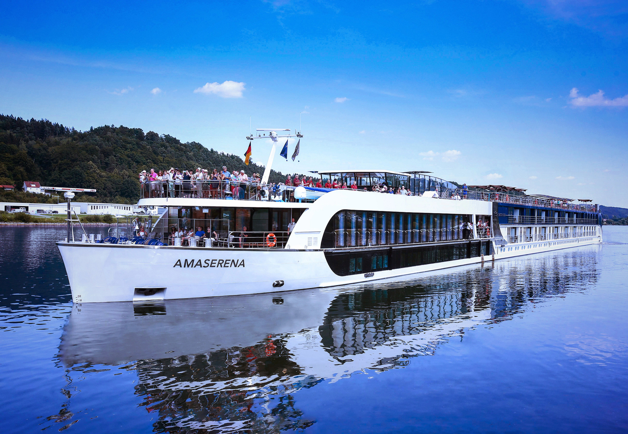 A modern white river cruise ship named AmaSerena docked on a serene blue river, with passengers visible on multiple decks against a backdrop of forested shoreline and blue sky.