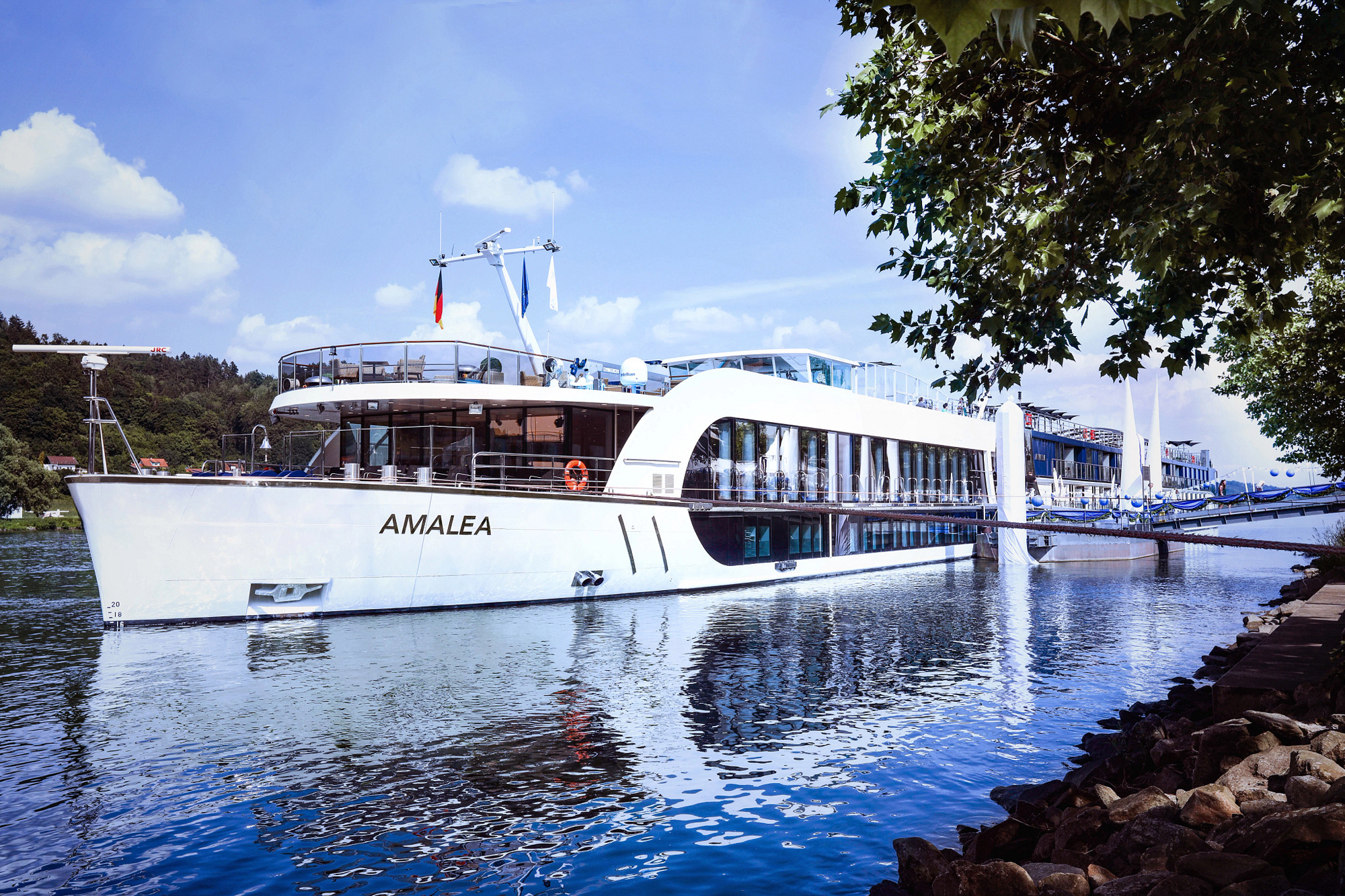 A modern white river cruise ship named Amalea moored alongside a rocky shoreline, with lush trees and blue sky in the background. The sleek vessel features large windows and multiple decks.