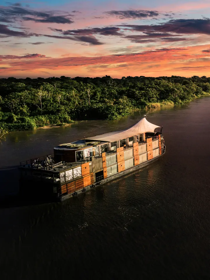 Amazon River Cruise at Sunset A traditional wooden riverboat cruise vessel navigates the Amazon River during a stunning golden sunset, with dense tropical rainforest vegetation lining both banks. The ship features a distinctive canvas canopy top and wooden construction, exemplifying authentic Amazonian river exploration.