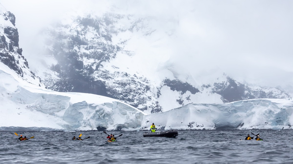 Antarctic Expedition Zodiac Adventure Adventure travelers in inflatable Zodiac boats explore the pristine Antarctic landscape, navigating through icy waters with massive snow-covered mountains and glaciers towering in the background under cloudy skies.