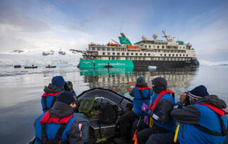 A group of travelers in blue and red waterproof gear ride a Zodiac boat near the Aurora Expeditions cruise ship in an icy Antarctic landscape with glaciers and snow-covered mountains in the background.