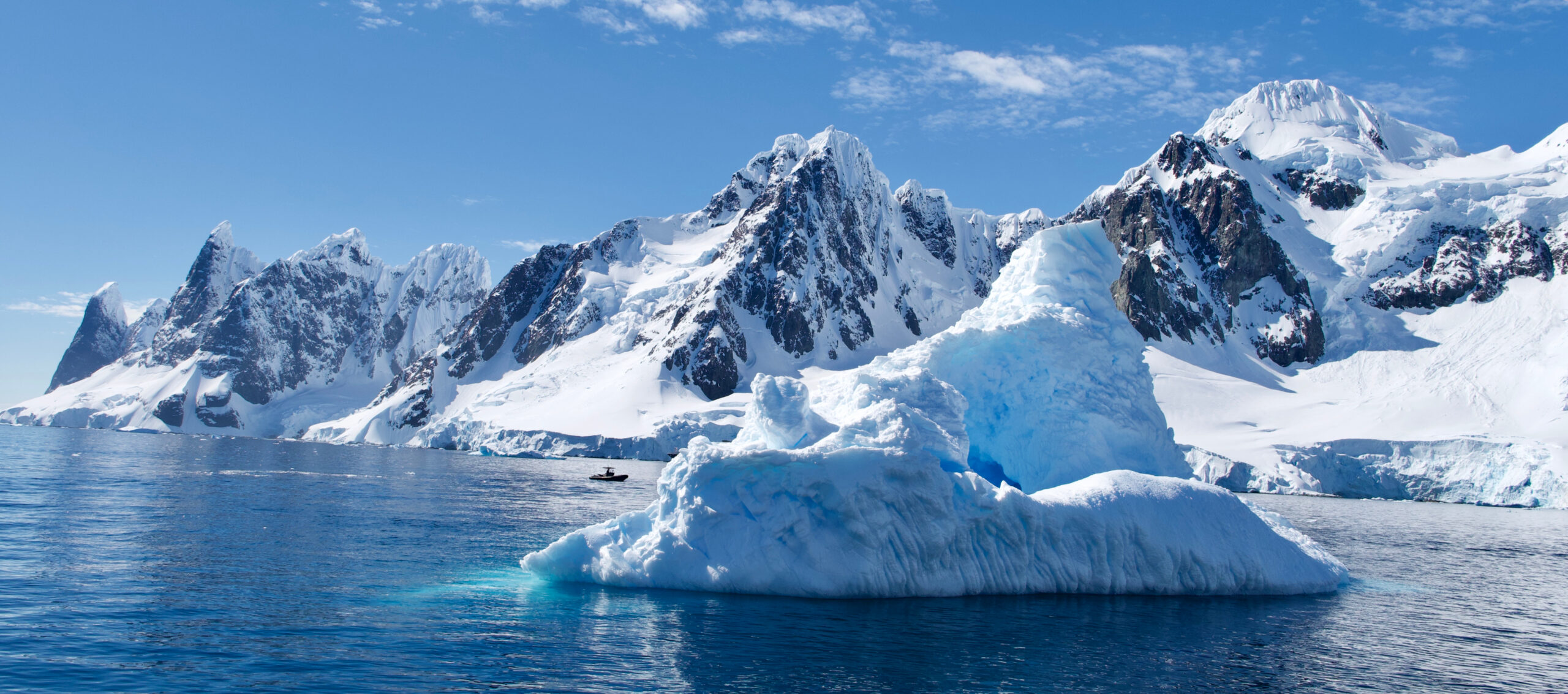 Antarctic Iceberg and Snowy Mountain Landscape A massive blue-white iceberg floating in the foreground with dramatic snow-covered mountains and rocky peaks in the background. A small boat can be seen navigating the pristine polar waters.