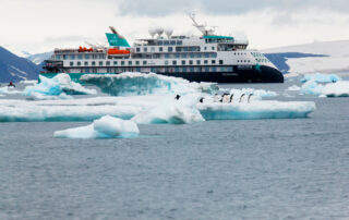 An Aurora Expeditions cruise ship navigating through icy waters with blue-white icebergs and a group of penguins on an ice floe in the foreground. Snowy mountains are visible in the background.
