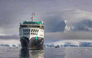 A white and teal expedition cruise ship navigating through icy Antarctic waters, with snow-covered mountains and glaciers in the background and floating ice fragments around the vessel.