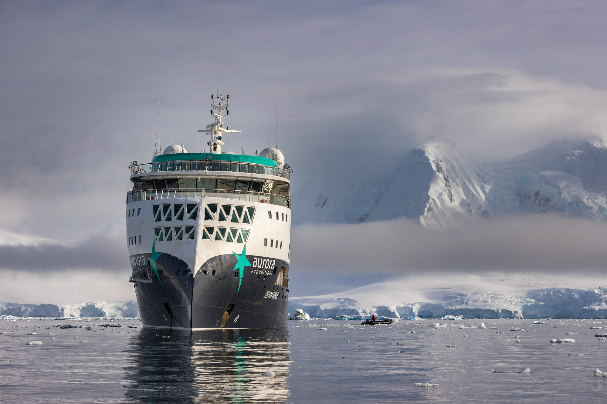 A white and teal expedition cruise ship navigating through icy Antarctic waters, with snow-covered mountains and glaciers in the background and floating ice fragments around the vessel.