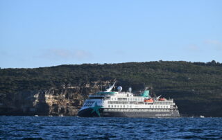An Aurora Expeditions expedition cruise ship sailing close to a rugged, rocky coastline with dense green vegetation on top of dramatic cliff formations. The ship features a distinctive teal and gray color scheme.