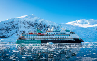 An expedition cruise ship navigating through icy waters with snow-capped mountains and glaciers in the background. The Aurora Expeditions' Sylvia Earle vessel is surrounded by floating ice in a pristine polar landscape.