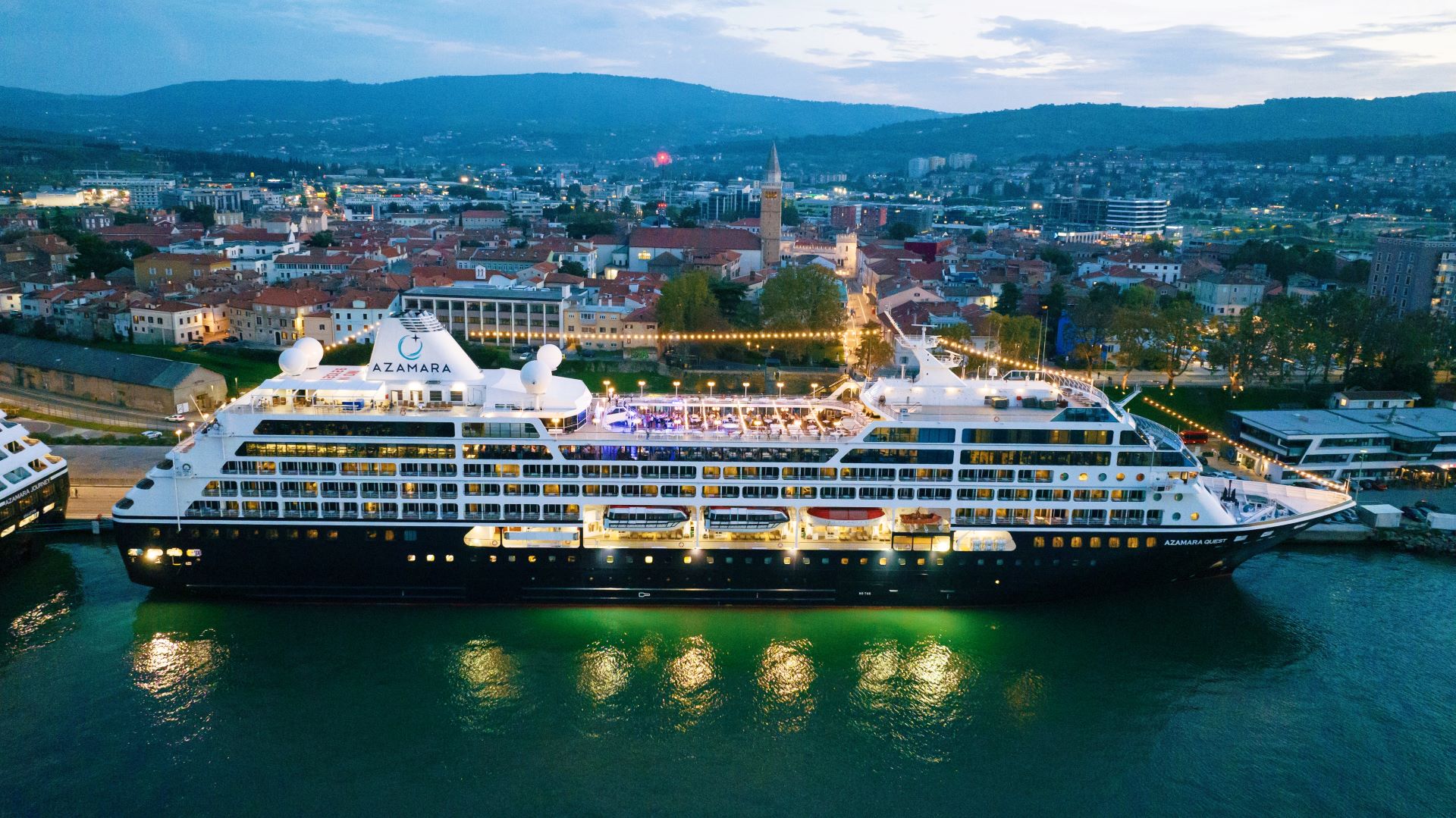 An Azamara cruise ship illuminated at night, docked in a picturesque European city with mountains and urban landscape in the background. The ship's lights reflect beautifully on the water, creating a stunning evening scene.
