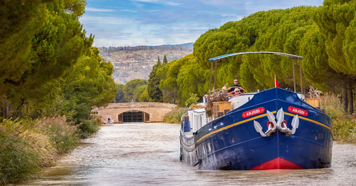 Barge Cruising Through European Canal Lock A traditional blue and gold barge named Anjobi navigates through a historic stone canal lock in a verdant European waterway, lined with tall cypress trees and framed by distant hillside settlements, showcasing scenic inland waterway tourism.