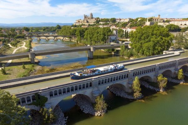 Barge Traversing Ancient French Waterway A modern river barge travels along the Lot River in southwestern France, passing beneath a historic multi-arched stone bridge with a medieval cathedral and fortified town visible on the hilltop in the background, showcasing Europe's rich cultural heritage and scenic waterway tourism.