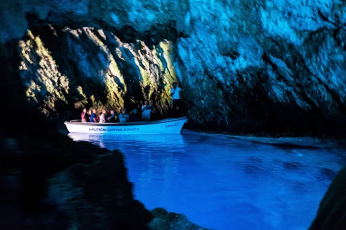 Blue Grotto Cave Boat Tour A stunning white multi-deck motor yacht cruises through crystal-clear Mediterranean waters with a picturesque coastal town featuring white buildings nestled against rocky hills in the background. The vessel represents premium yacht charter and luxury cruise travel experiences in Mediterranean destinations.