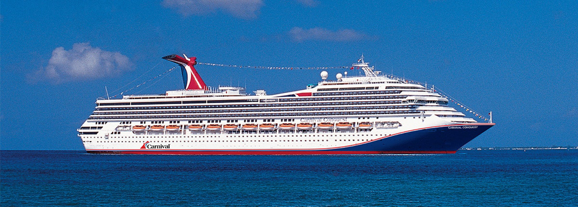 A large white and red Carnival cruise ship sailing on deep blue ocean waters under a clear blue sky with soft clouds. The vessel features multiple decks and an distinctive red funnel, representing typical modern cruise ship design.