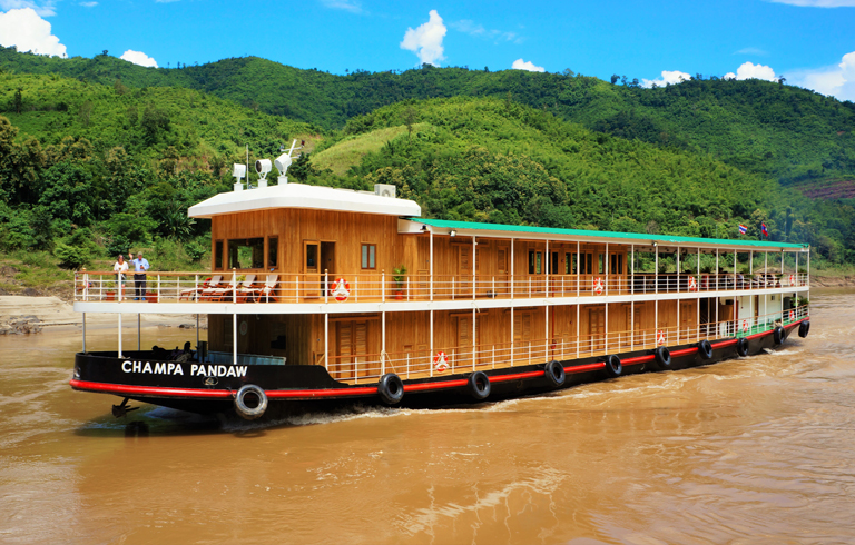 Champa Pandaw River Cruise in Lush Landscape A wooden river cruise boat named Champa Pandaw sailing on a muddy river with green mountainous terrain in the background. The vessel appears to be a traditional style river cruiser with multiple decks and wooden construction.