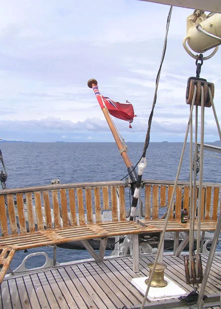 A traditional wooden sailing ship's deck is captured from an elevated vantage point, featuring a red flag mounted on the wooden railings, maritime rigging equipment, and pulleys against a calm sea and overcast sky. The image showcases the classic nautical heritage and sailing traditions with detailed views of the ship's wooden deck infrastructure and traditional maritime equipment.