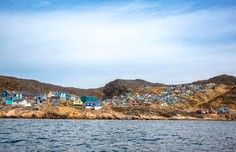 Coastal Village Harbor View A picturesque coastal settlement nestled against arid, rocky hills viewed from the water, featuring colorful buildings clustered along the shoreline with a calm sea in the foreground and blue sky above. The architecture and landscape suggest a Mediterranean or Middle Eastern location with a dry, semi-arid climate.