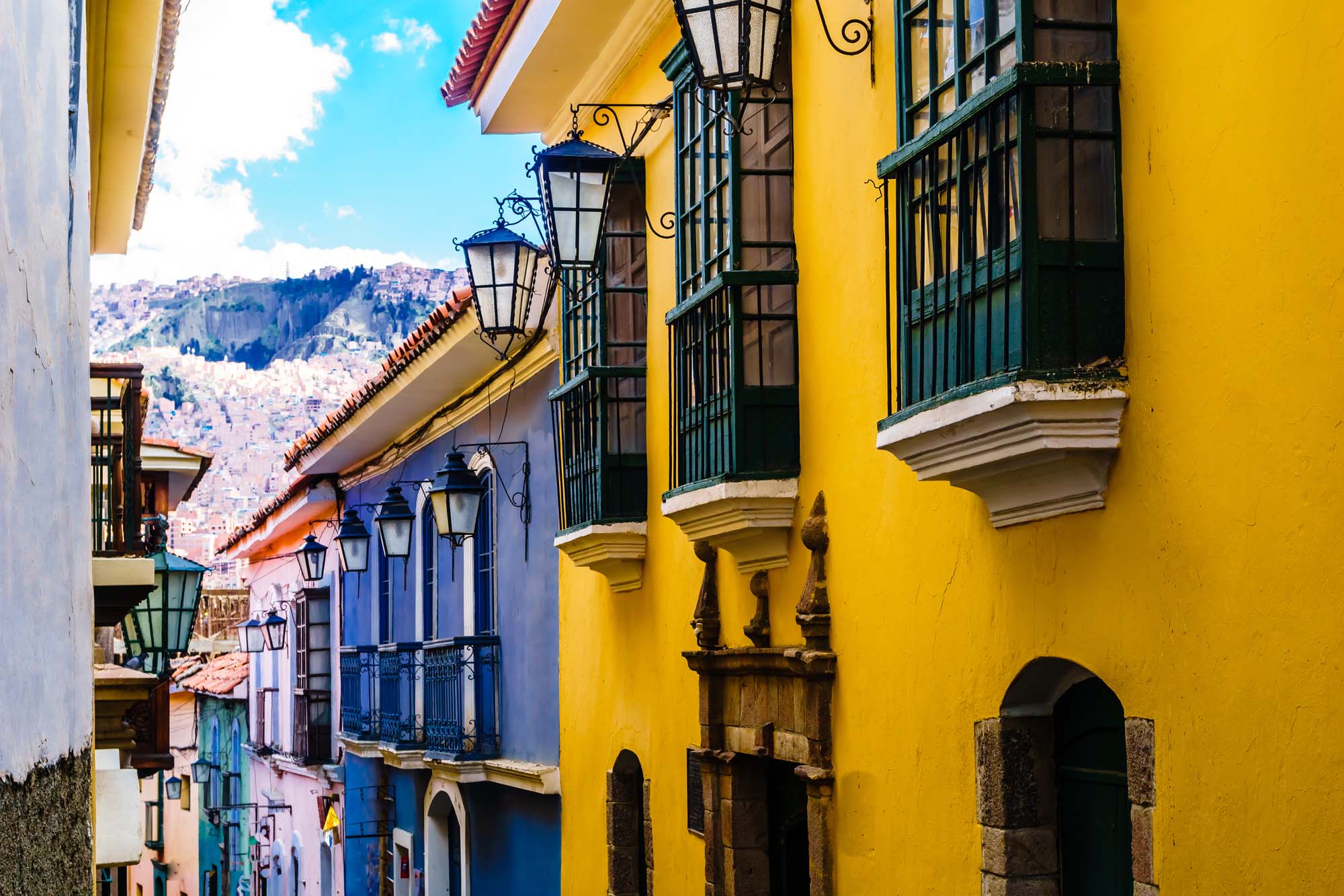 A vibrant street scene showcasing colonial architecture with bright yellow and blue buildings, ornate balconies, and traditional street lamps, set against a mountainous backdrop.