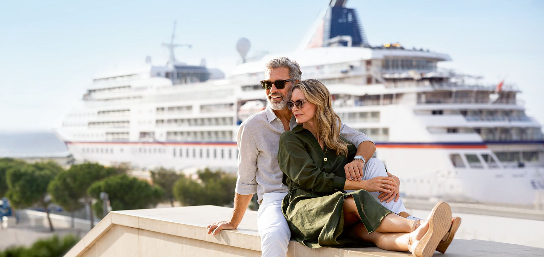 Couple Enjoying Cruise Ship View A mature couple sits together on a waterfront ledge, gazing at a large cruise ship docked in the harbor behind them. Both wear sunglasses and casual summer clothing, capturing a romantic moment during their cruise vacation with the impressive vessel and coastal scenery in the background.