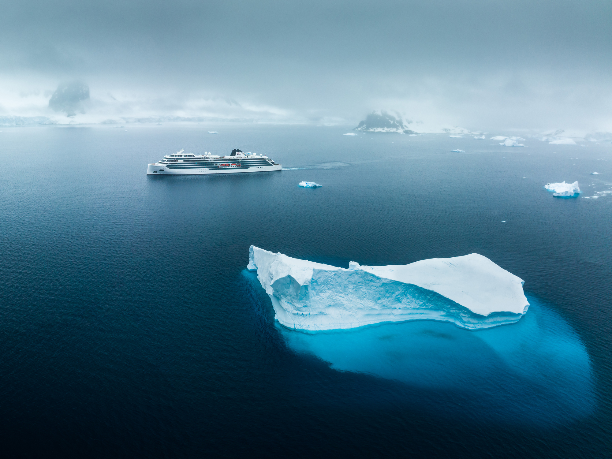 Cruise Ship Amidst Polar Icebergs A modern white cruise ship sailing through cold arctic waters near large, blue-white icebergs with misty mountainous islands in the background. The image captures the serene and dramatic landscape of a polar expedition.