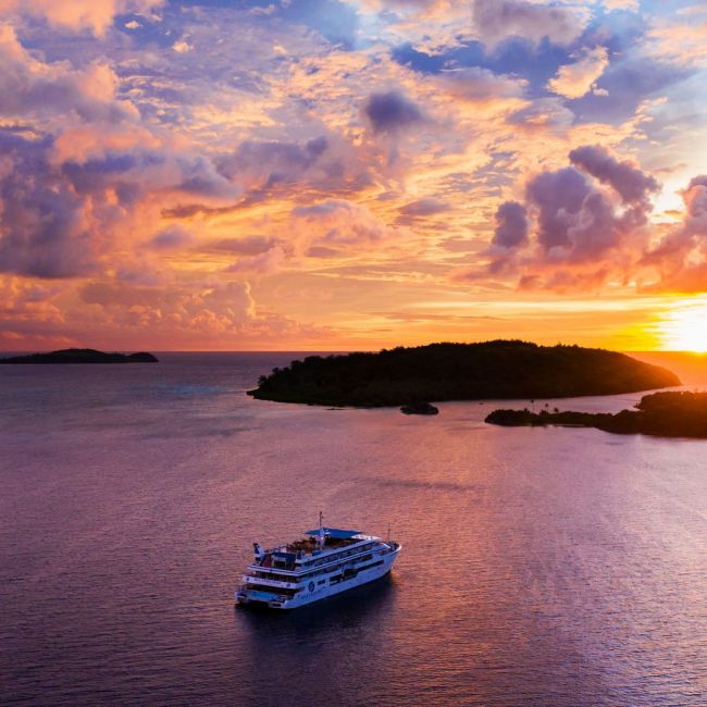 Cruise Ship Anchored at Tropical Sunset A modern cruise ship is anchored in calm turquoise waters during a spectacular golden and purple sunset, with a lush forested island silhouetted in the background and dramatic cloud formations illuminated by the setting sun.