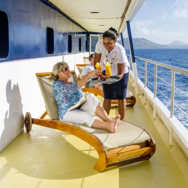 Cruise Ship Deck Service with Ocean View A crew member serves refreshments to a passenger relaxing on a lounge chair on the promenade deck of a cruise ship, with panoramic ocean views and volcanic islands visible on the horizon.