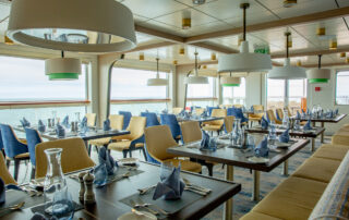 An elegant dining area on a cruise ship featuring modern decor with blue and yellow chairs, large windows overlooking the sea, and tables set with white tablecloths and blue napkins.