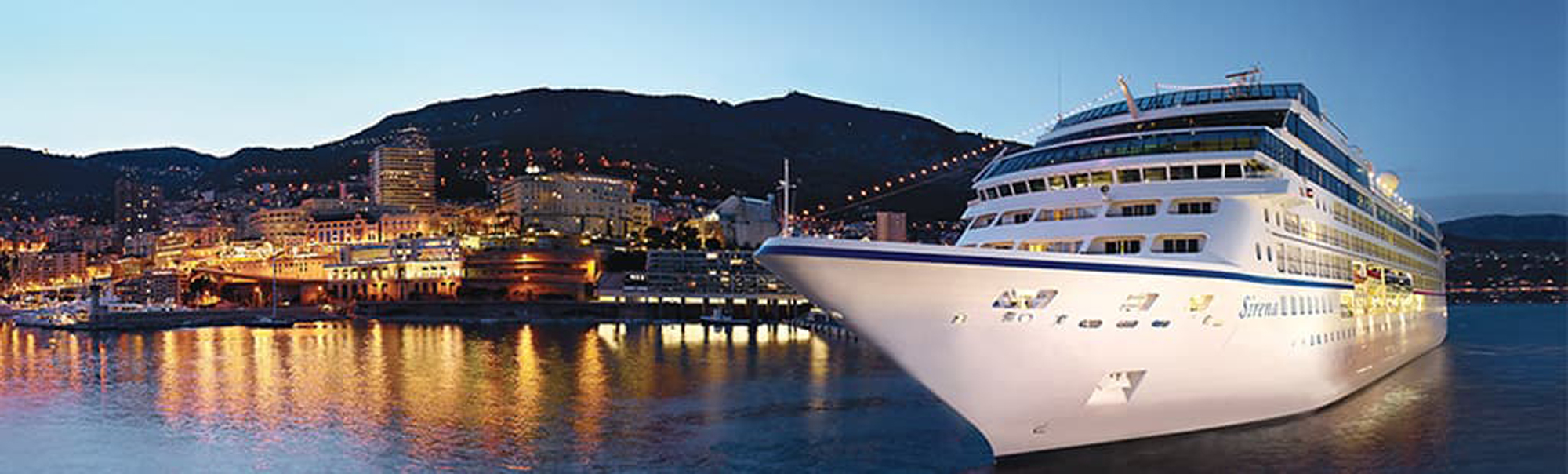 Cruise Ship Docked in Coastal City at Night A large white cruise ship moored at a vibrant nighttime port with illuminated city buildings and mountainous backdrop. The ship's lights reflect on the calm water, creating a picturesque maritime scene.