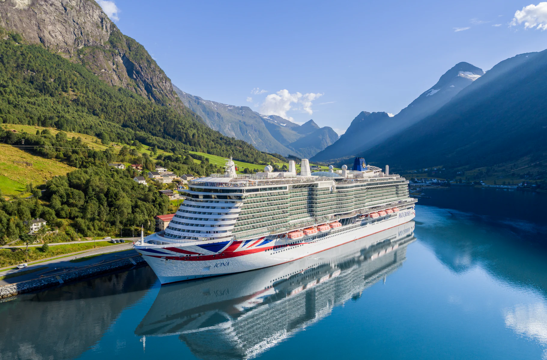 A large modern cruise ship is moored in a pristine Norwegian fjord surrounded by dramatic steep mountain cliffs, lush green forests, and crystal-clear turquoise waters. The picturesque alpine valley setting showcases the natural beauty of Scandinavia with snow-capped peaks in the distance and traditional Norwegian architecture visible on the shoreline.