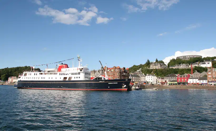 Cruise Ship Docked in Oban Harbor A large passenger cruise ship with distinctive red and white livery is moored in the picturesque Scottish port town of Oban, with colorful waterfront buildings and a hilltop structure visible in the background under clear blue skies.
