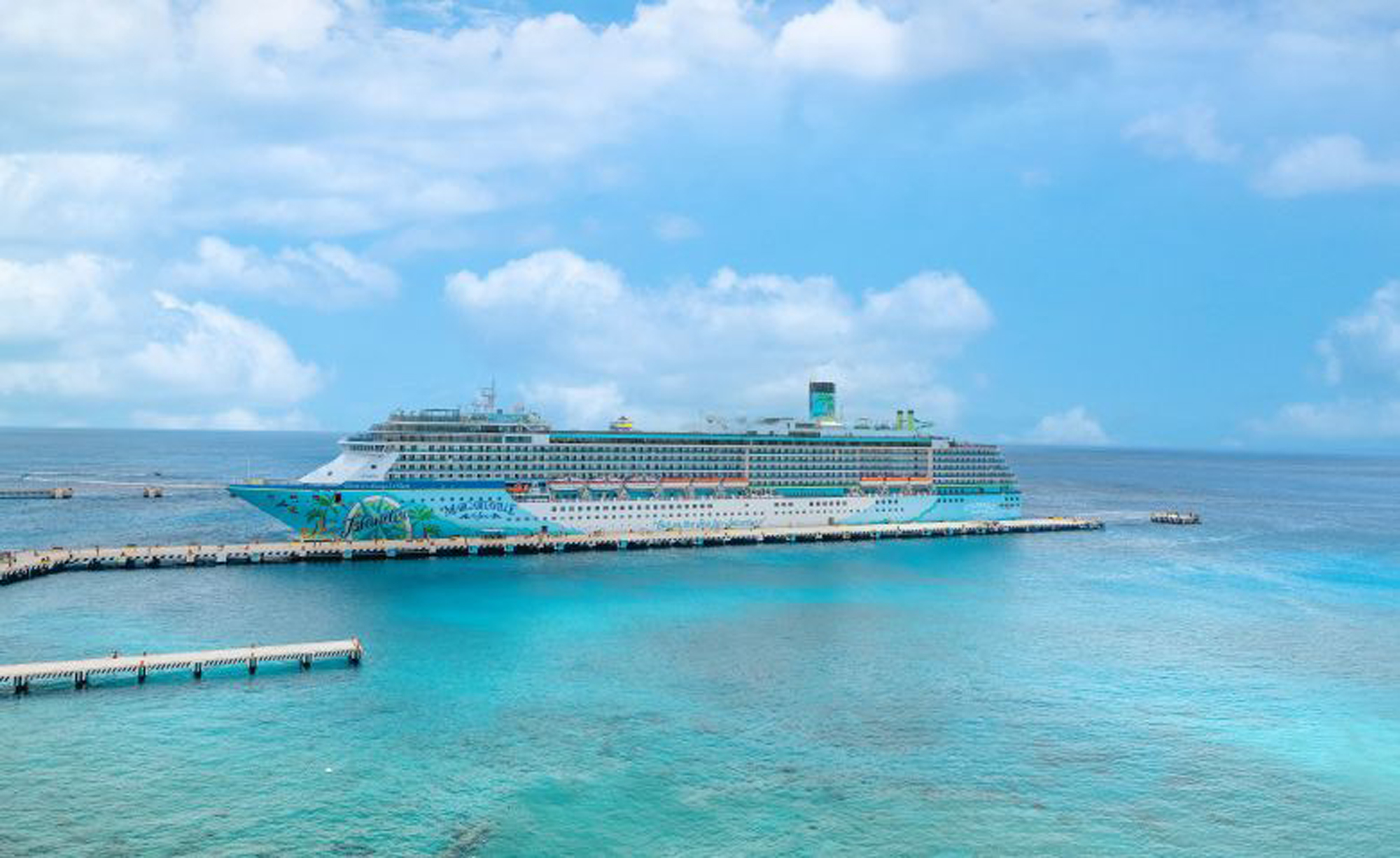 Cruise Ship Docked in Turquoise Caribbean Waters A large, colorfully decorated cruise ship moored at a pier with crystal clear blue-green waters surrounding it. The ship is set against a bright blue sky with fluffy white clouds.