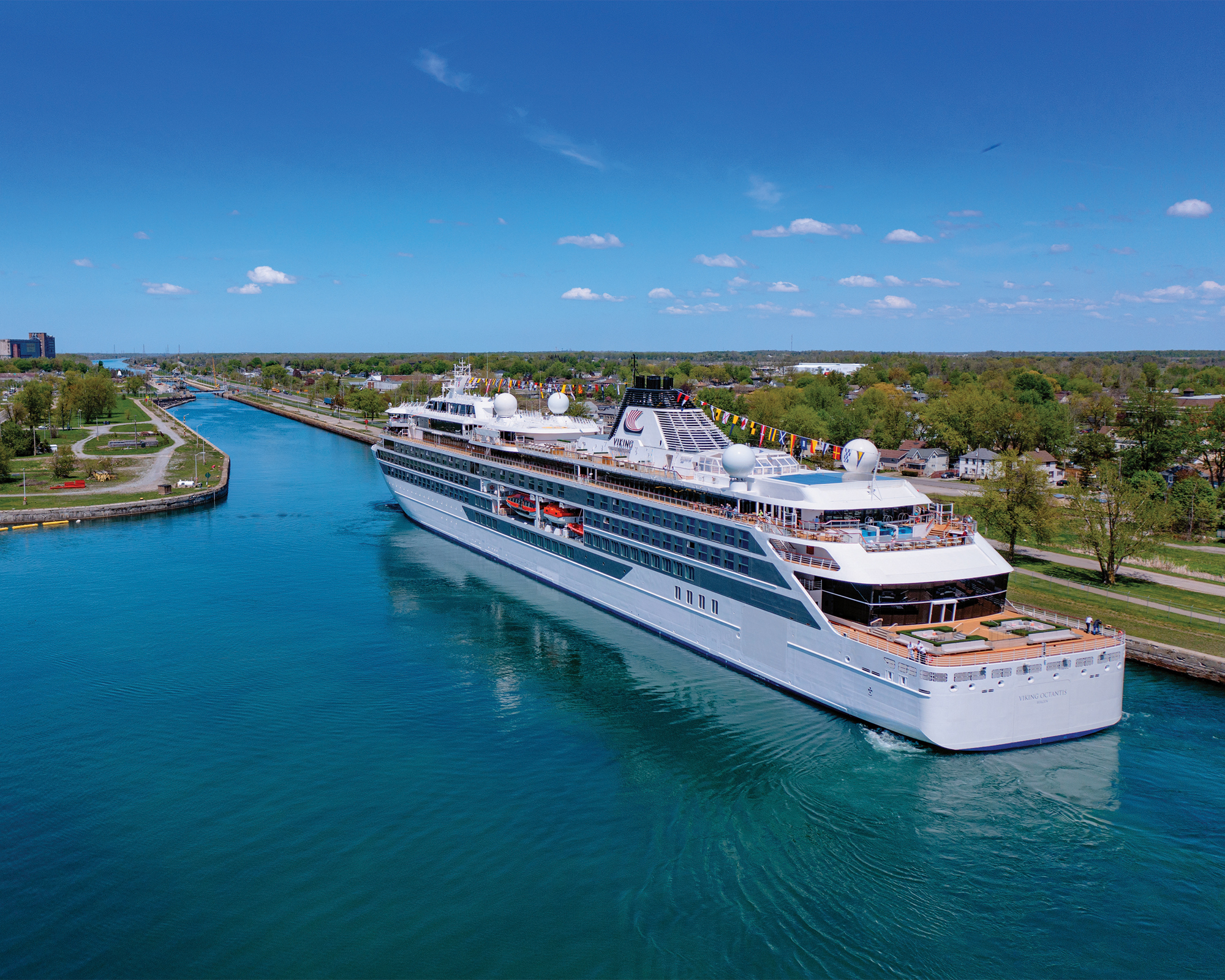 Cruise Ship Navigating Scenic Waterway A sleek white cruise ship sailing through a bright blue canal with lush green shoreline and urban landscape in the background. The vessel is adorned with colorful flags and appears to be in pristine condition.