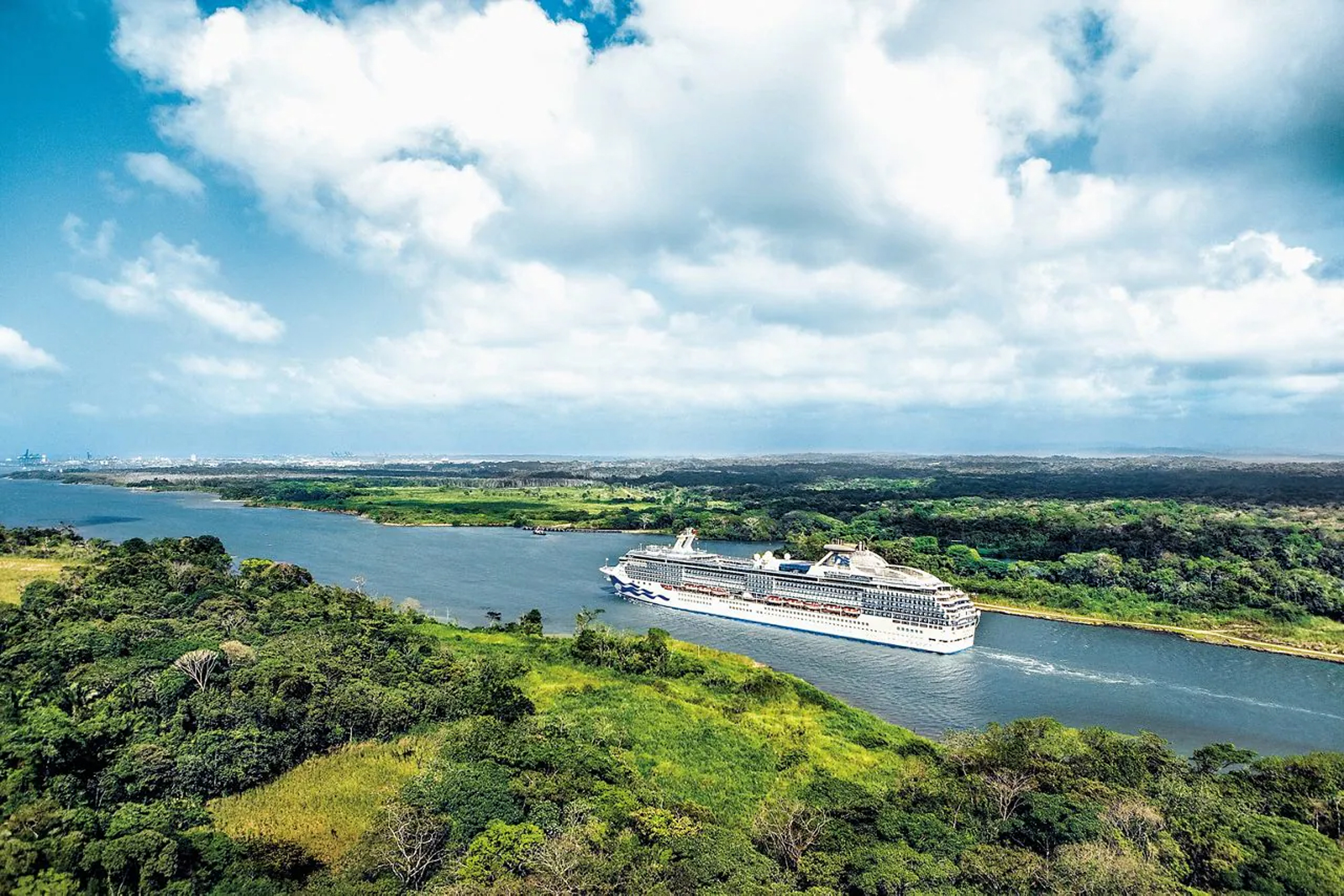 A large white cruise ship sailing through a lush green landscape with dense forests and blue waters. The ship is framed by verdant coastlines under a bright, cloudy sky.