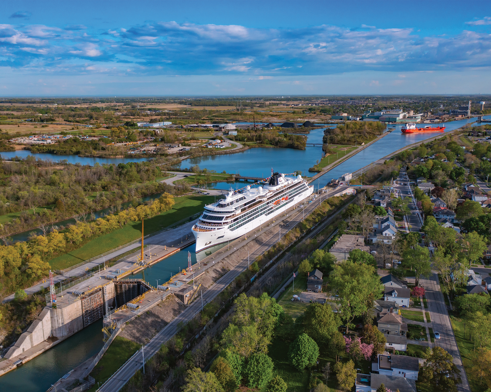 Cruise Ship Passing Through Canal Lock An aerial view of a large white cruise ship navigating through a narrow canal lock, surrounded by green landscapes and residential areas on a clear blue sky day.