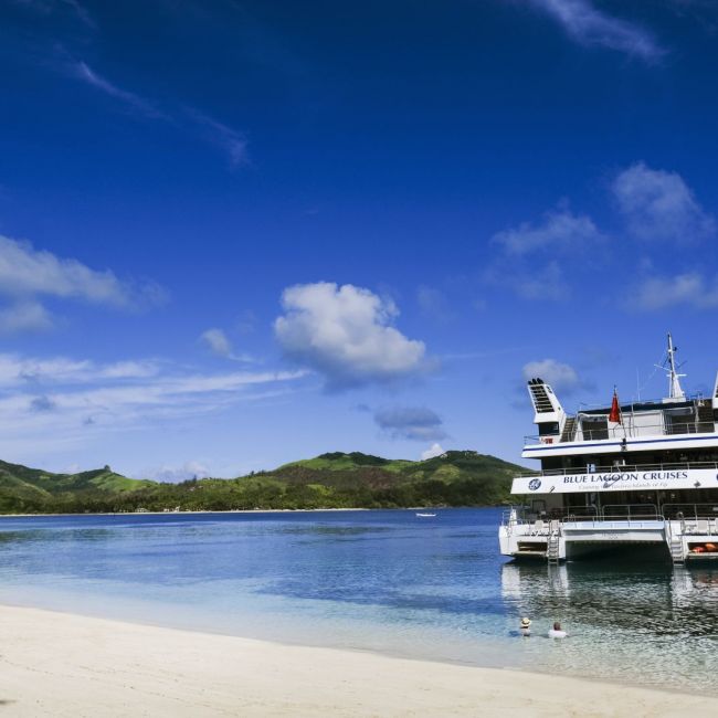 Cruise Ship at Tropical Island Beach A Holland America Line cruise ship is docked at a pristine tropical beach with turquoise waters, white sand, and lush green volcanic mountains in the background under a brilliant blue sky with white clouds.