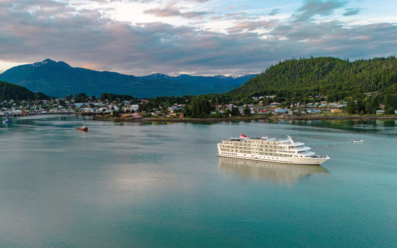 Cruise Ship in Alaskan Coastal Town A white cruise ship anchored in a tranquil bay near a forested coastal town with mountains in the background. The calm waters reflect the cloudy sky and surrounding landscape.