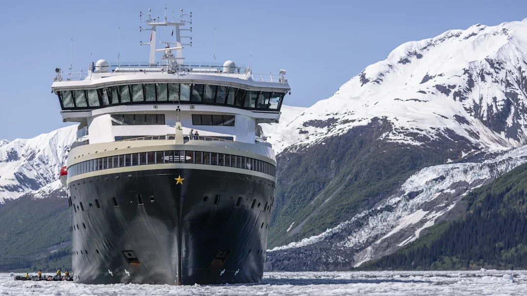 A large cruise ship sailing through icy waters with snow-capped mountains and glacial terrain in the background. A small zodiac boat is visible near the ship's hull.