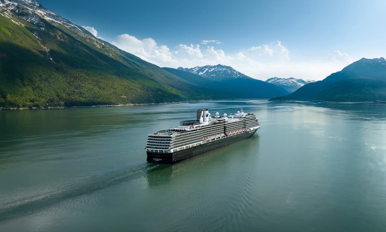 Cruise Ship in Alaskan Mountain Fjord A modern cruise ship sailing through a serene fjord surrounded by lush green mountains and snow-capped peaks under a clear blue sky. The vessel appears to be navigating through calm, reflective waters with dramatic Alaskan wilderness in the background.