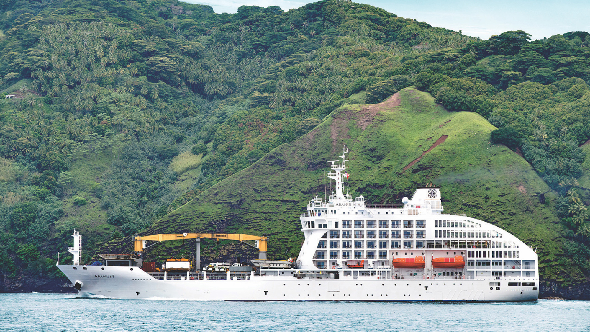 Cruise Ship in Lush Tropical Landscape A white cruise ship named Aranui 5 anchored near a densely forested mountainous coastline with green hills and tropical vegetation. The ship appears to be a specialized expedition or cargo-cruise vessel positioned in a scenic maritime setting.