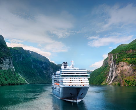 Cruise Ship in Norwegian Fjord A large cruise ship navigating through a dramatic fjord landscape with steep green mountains and a cascading waterfall. The vessel is surrounded by calm, blue-green waters and under a partly cloudy sky.