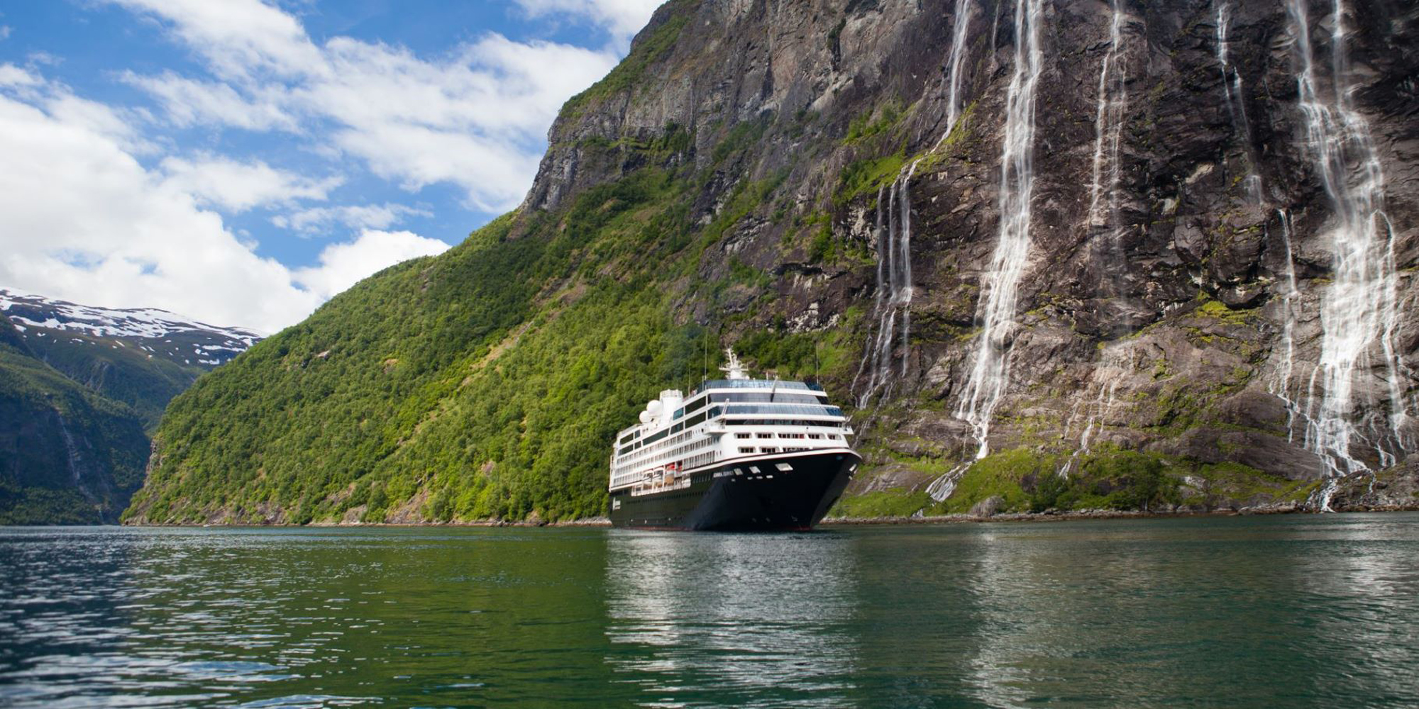 A white cruise ship sailing through a dramatic fjord landscape with steep, green-covered mountains and multiple waterfalls cascading down rocky cliffs. The calm, green-tinted water reflects the surrounding natural scenery.