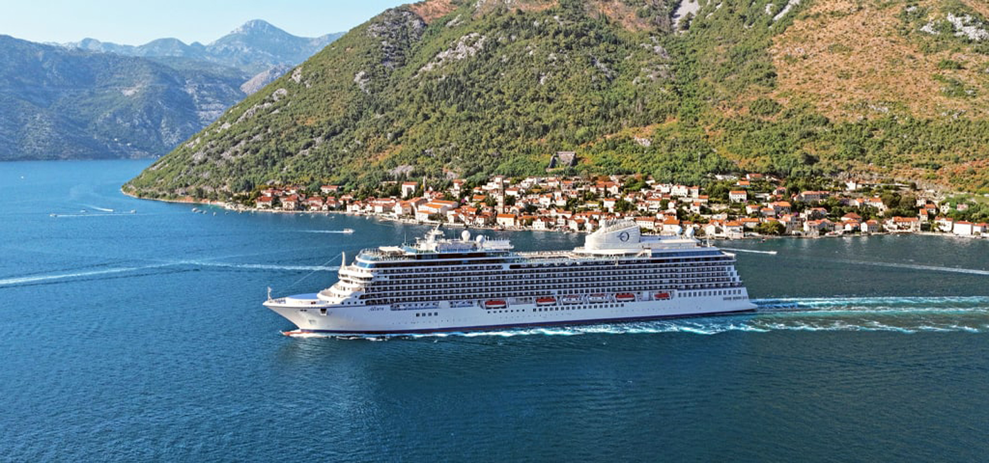 Cruise Ship in Scenic Coastal Mediterranean Town A large white cruise ship anchored near a picturesque coastal town nestled against mountainous terrain, with red-roofed buildings and blue waters creating a stunning Mediterranean landscape.