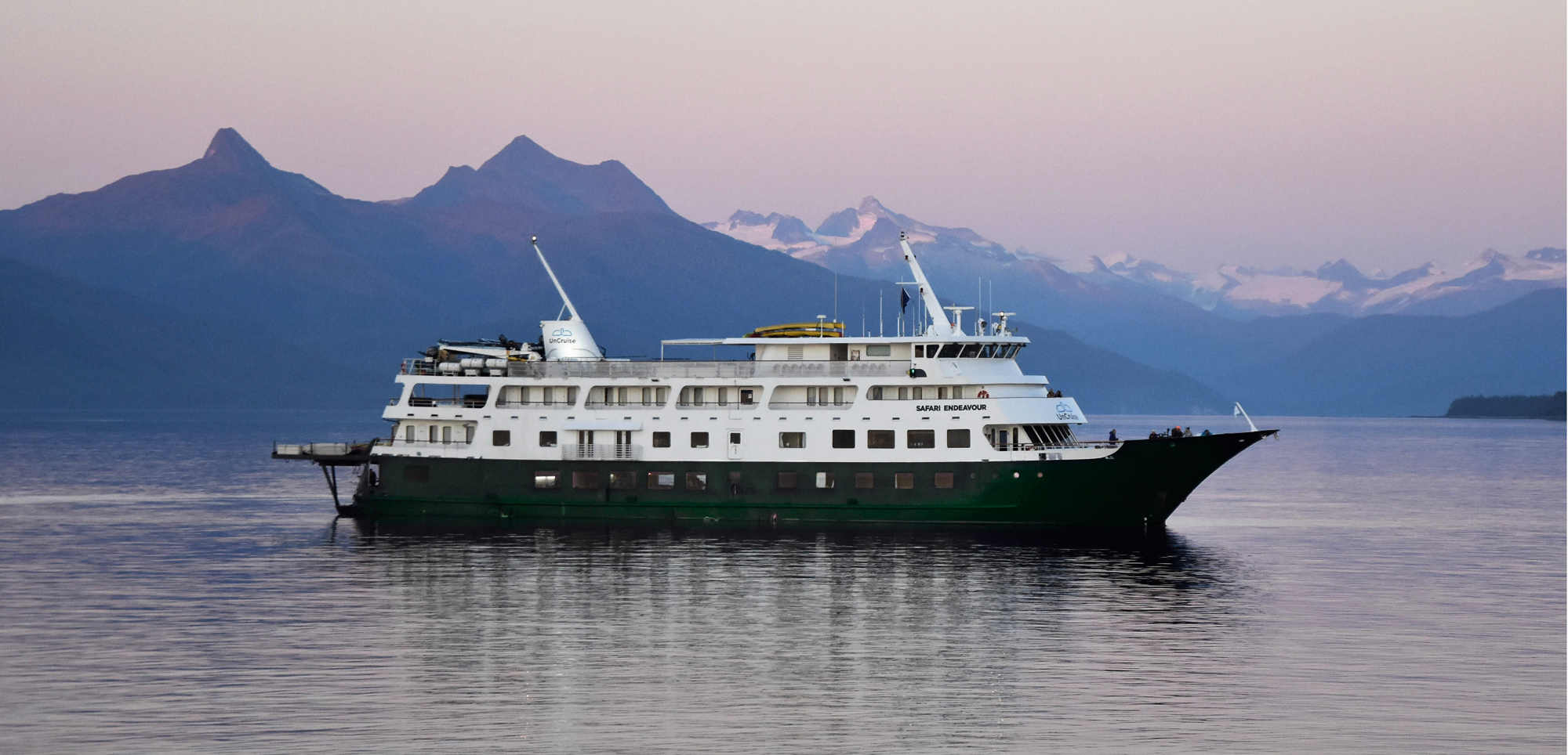 Cruise Ship in Scenic Mountain Fjord A white and green expedition cruise ship sailing on calm waters with dramatic mountain ranges and snow-capped peaks in the background during a soft-colored twilight or dawn.