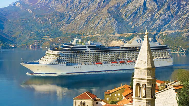 Cruise Ship in Scenic Mountain Fjord A large white cruise ship sailing through a stunning coastal landscape with dramatic mountain scenery and a charming historic town in the foreground. The vessel is anchored near a picturesque European coastal village with stone buildings and a church bell tower.